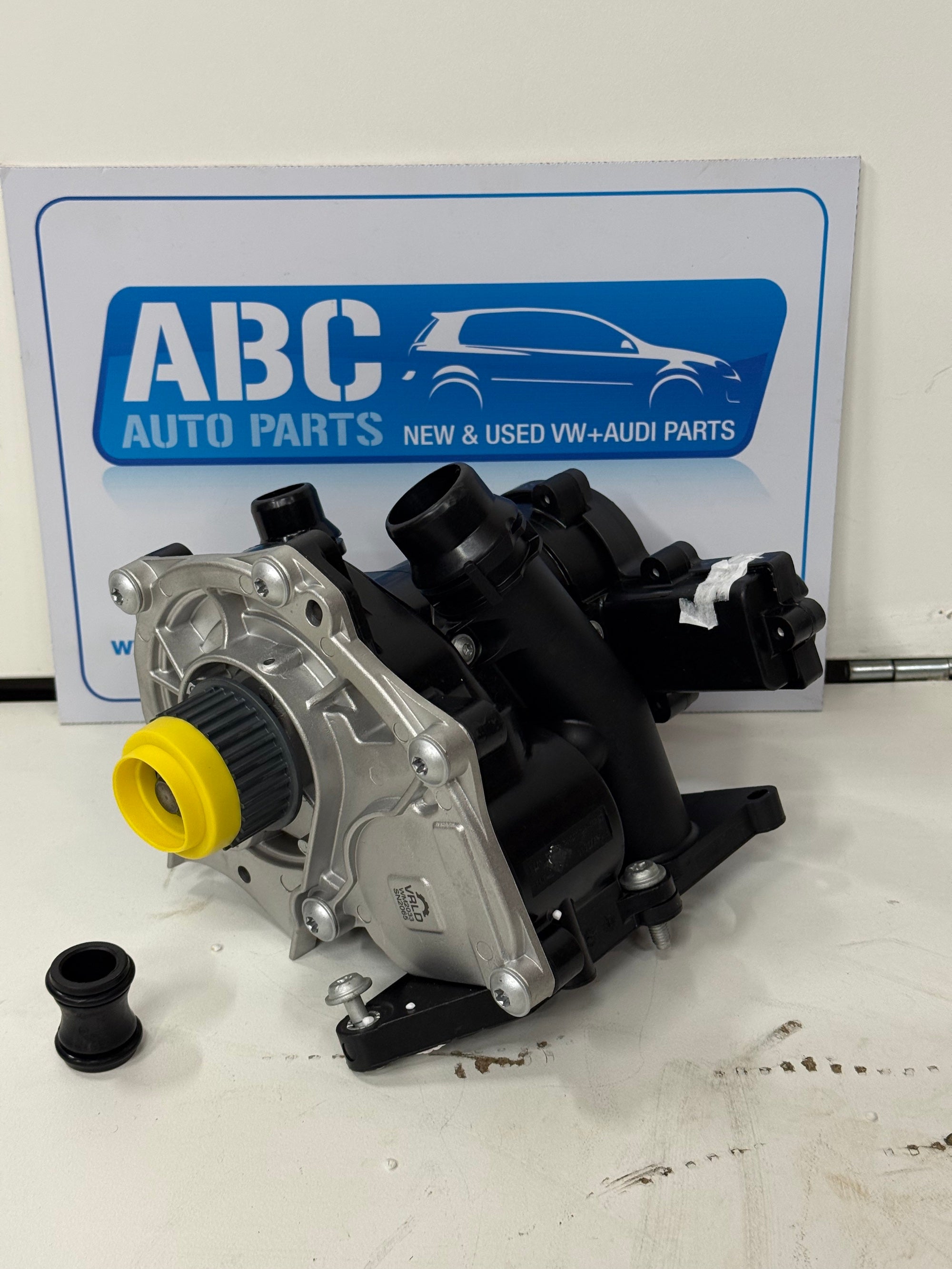 Car water pump with yellow seal and black parts on a white surface, with ABC Auto Parts sign in the background.